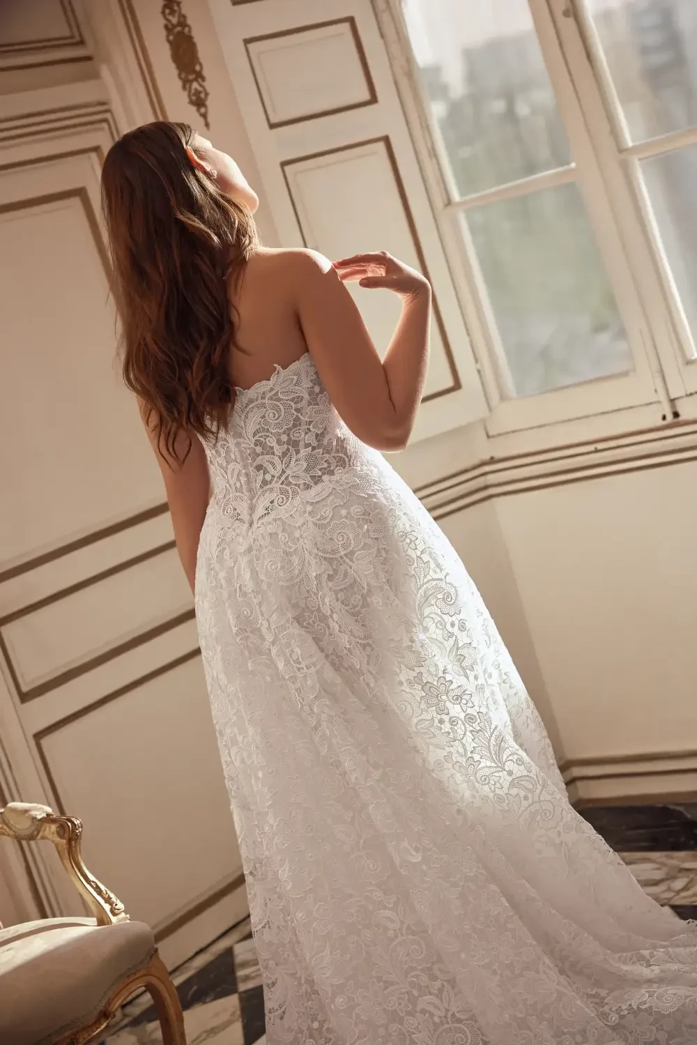 A woman in a white lace dress is standing with her back to the camera, gazing out of a window in a well-lit room.