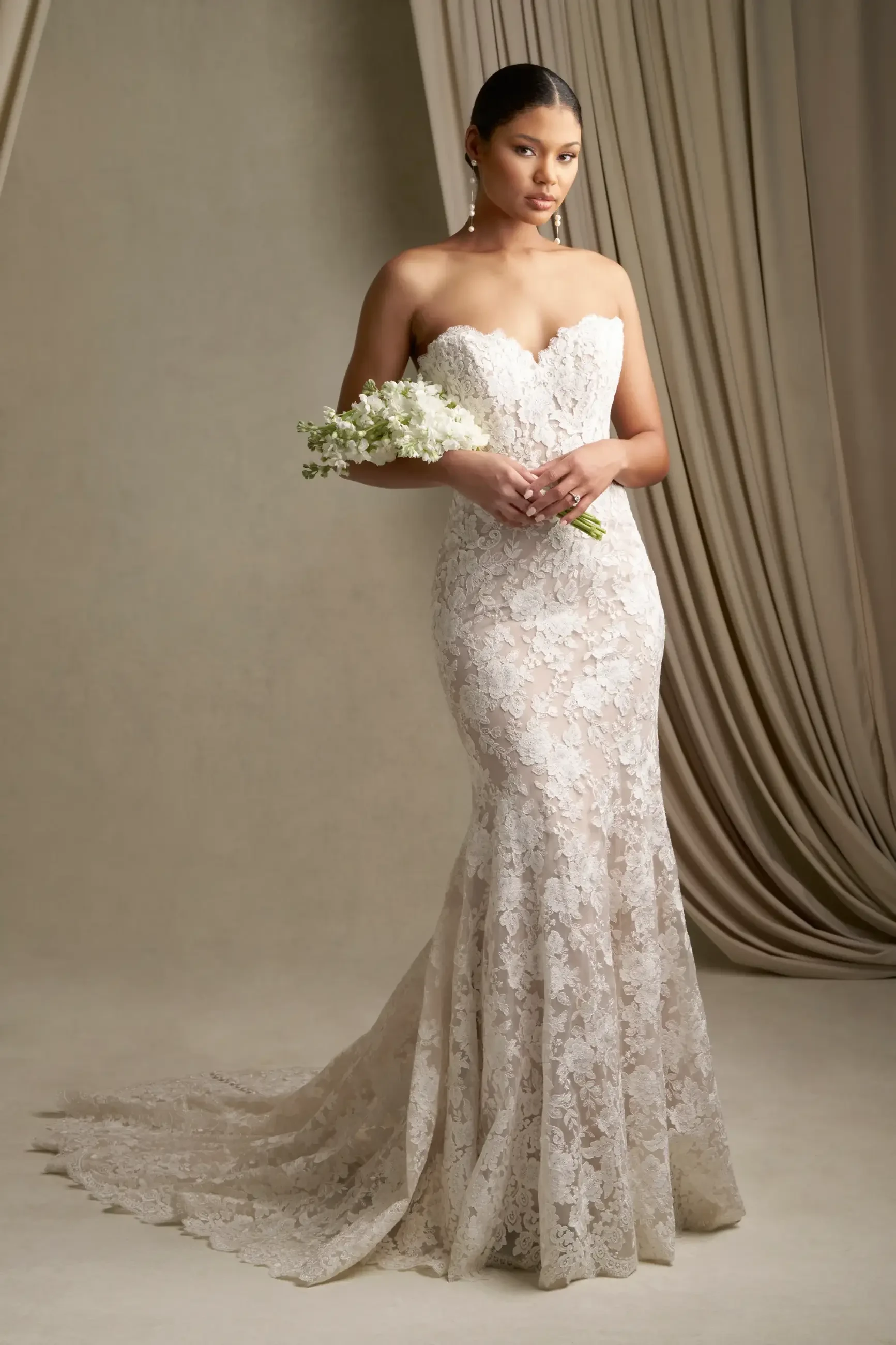 A woman in an elegant strapless lace wedding dress holds a small bouquet. She stands on a soft beige floor with flowing drapes, exuding a serene and poised demeanor.
