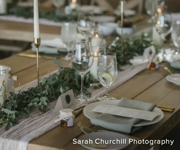A beautifully set dining table with candles, glasses of water, and wine, adorned with greenery and a menu on each plate.
