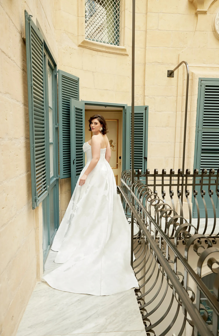 A woman in a white wedding dress standing on a balcony with blue shutters.