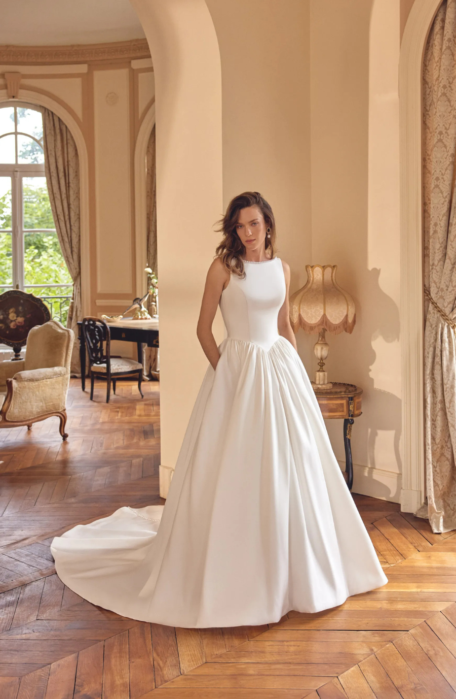 A woman in a simple white gown with pockets stands in a stylishly decorated room featuring large windows and elegant furnishings.